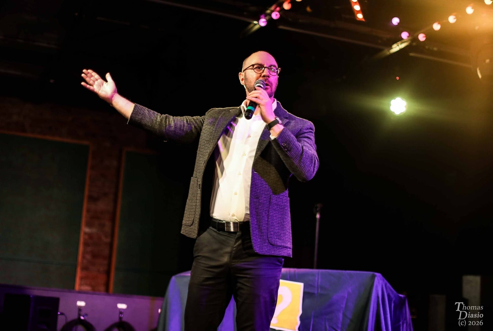 Max on stage with a microphone, arm outstretched toward the audience