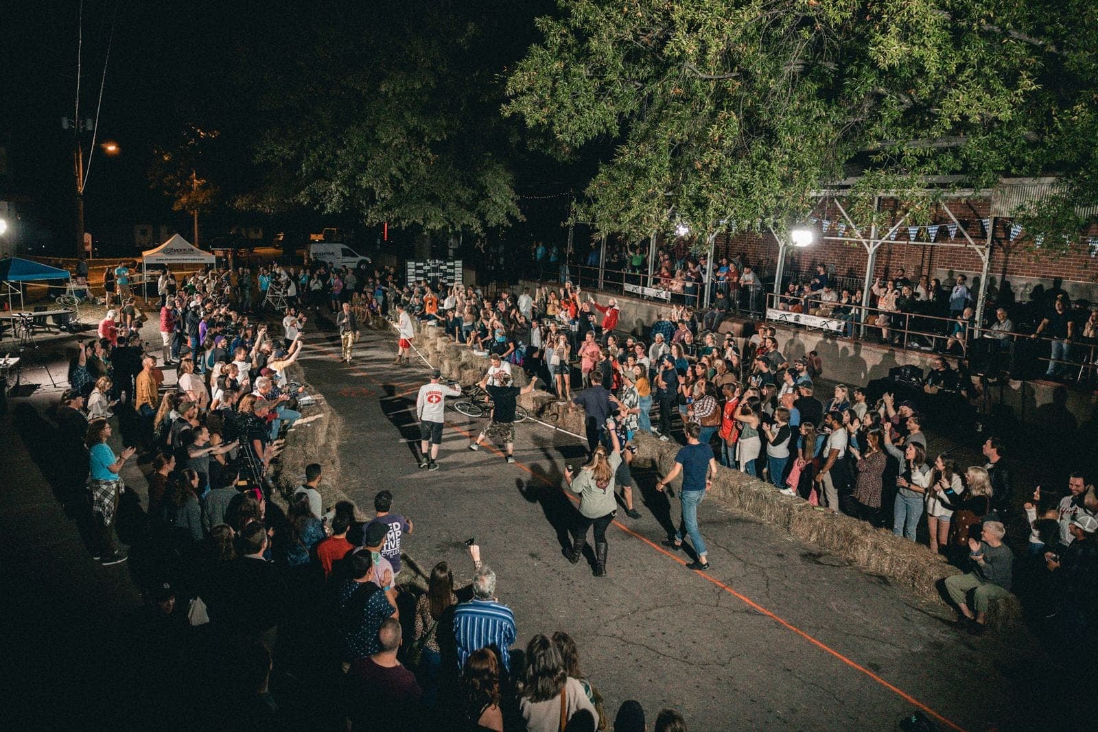 An aerial view of a huge nighttime crowd cheering around the hay-bale arena at the Tall Bike Joust