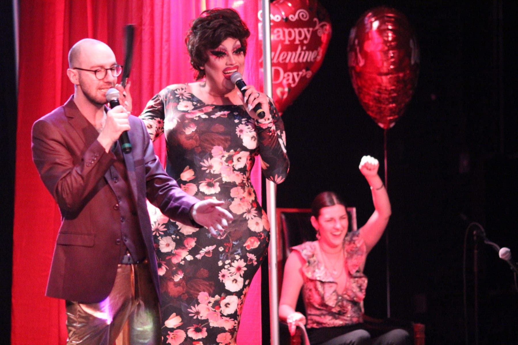 Max and his drag queen co-host on stage while a woman in the audience cheers