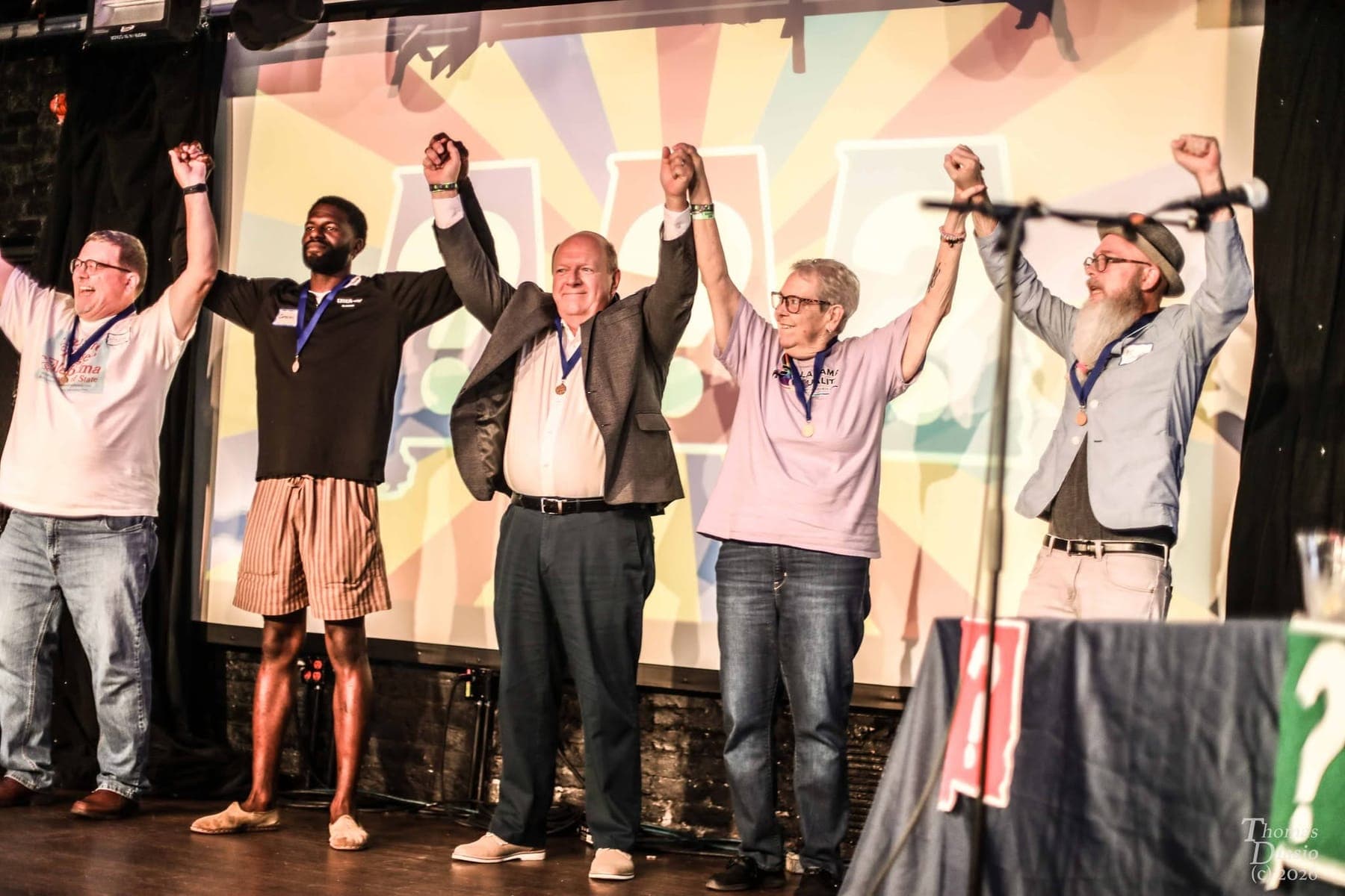 Five contestants wearing medals hold hands and raise their arms in celebration on stage