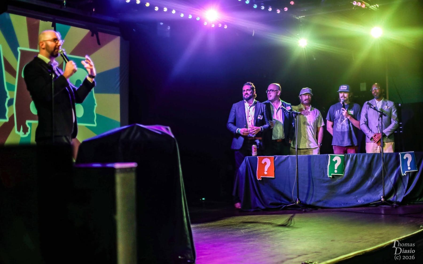 Max hosting while contestants stand behind game-show tables under colored stage lights